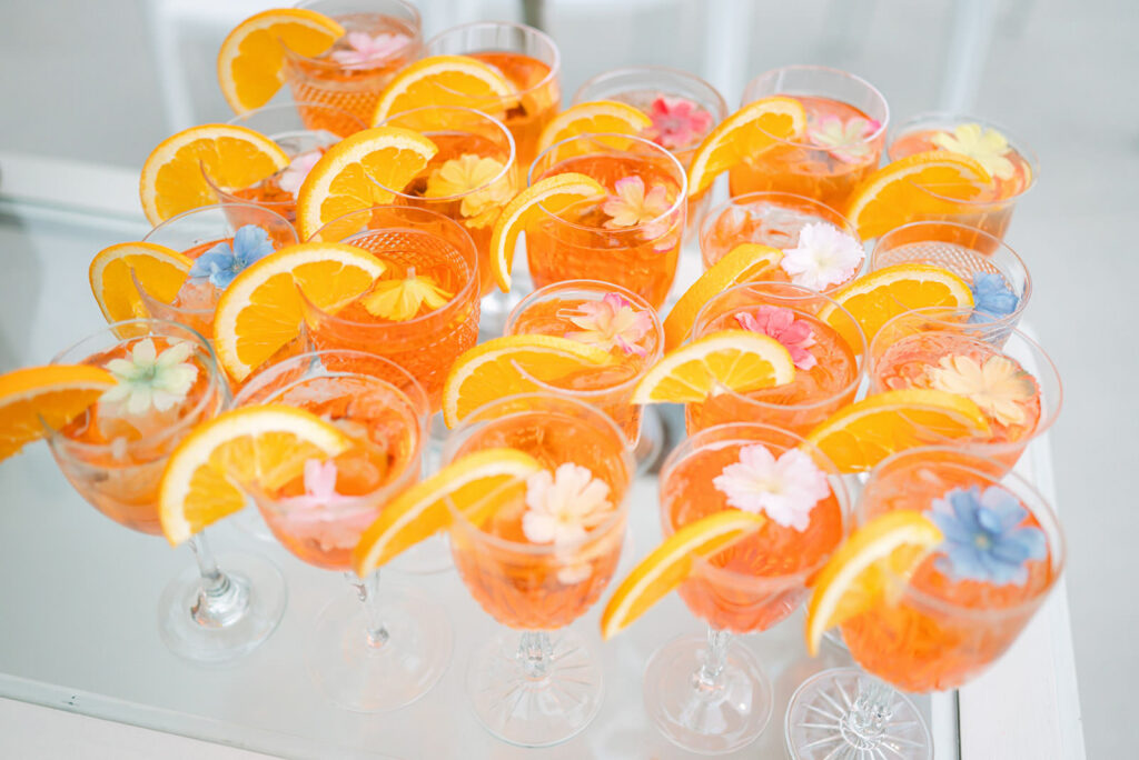 Summer Wedding Signature Drinks with Floral Garnish Orange signature wedding drinks in stemmed glasses with flowers floating on top, styled for a summer wedding celebration